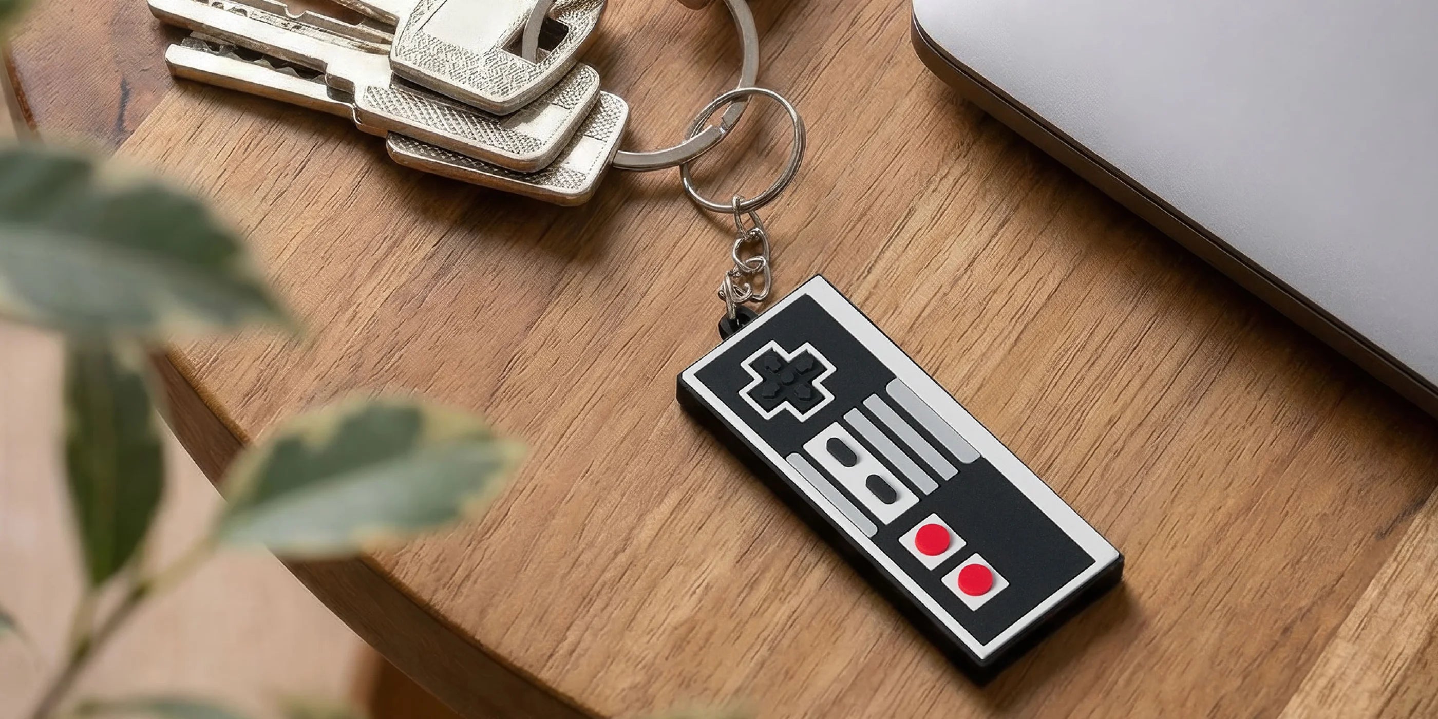 Keychain shaped like a classic gaming controller on a wooden surface with a plant and laptop in the background.